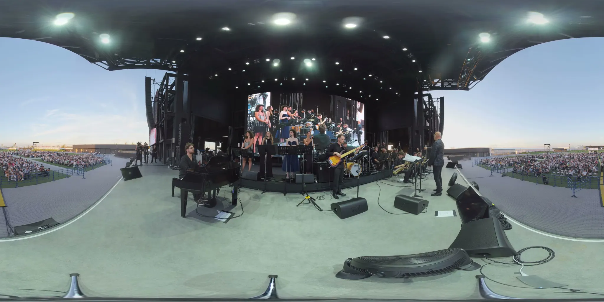 360° frame: a jazz orchestra on an open-air stage, singers at the centre, piano on the left, saxophones and brass on the right, the audience behind.