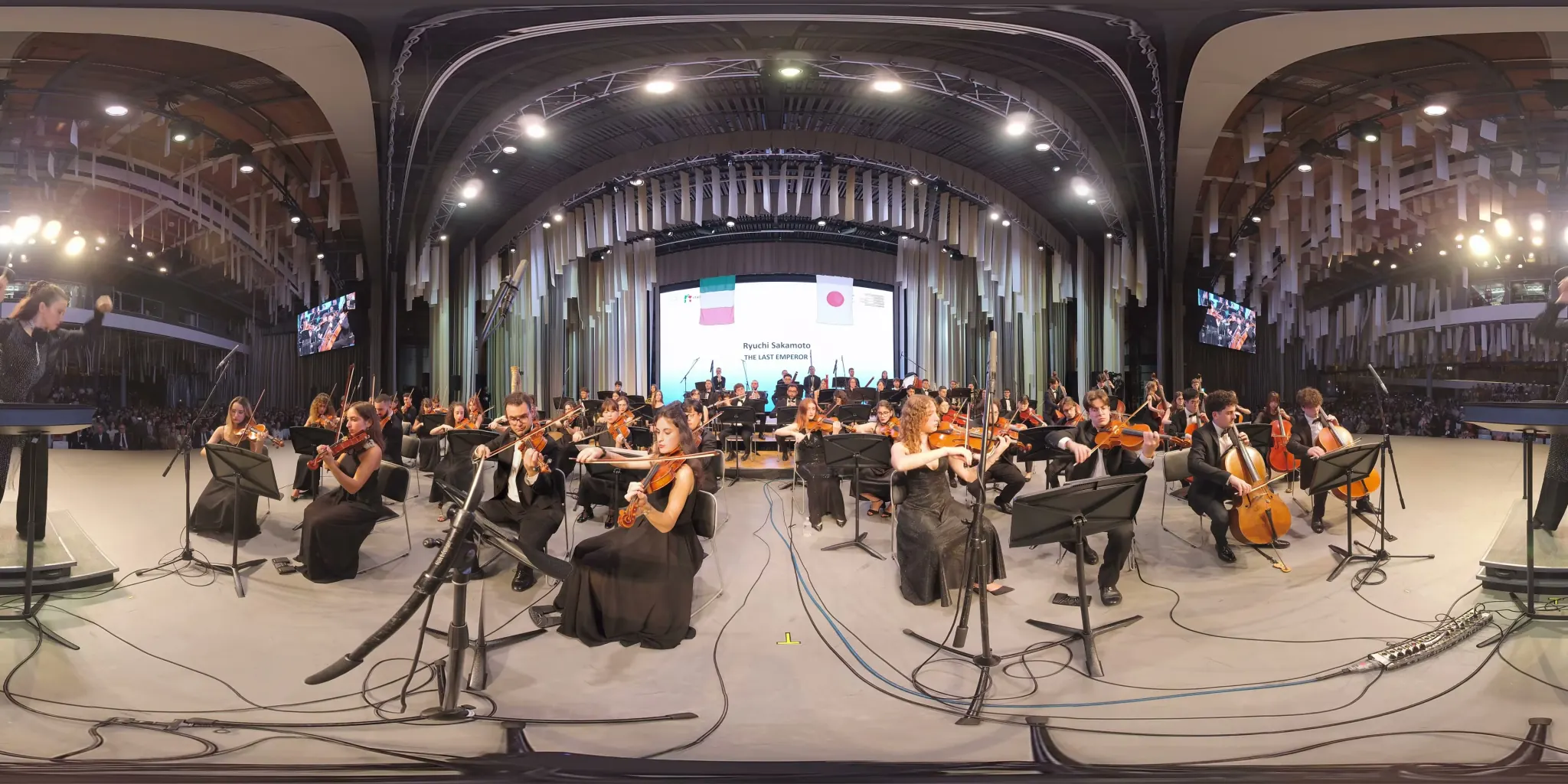 360° frame: a symphony orchestra seen from the podium, violins on the left, cellos on the right; on the screen at the back, the title “The Last Emperor” and the Italian and Japanese flags.